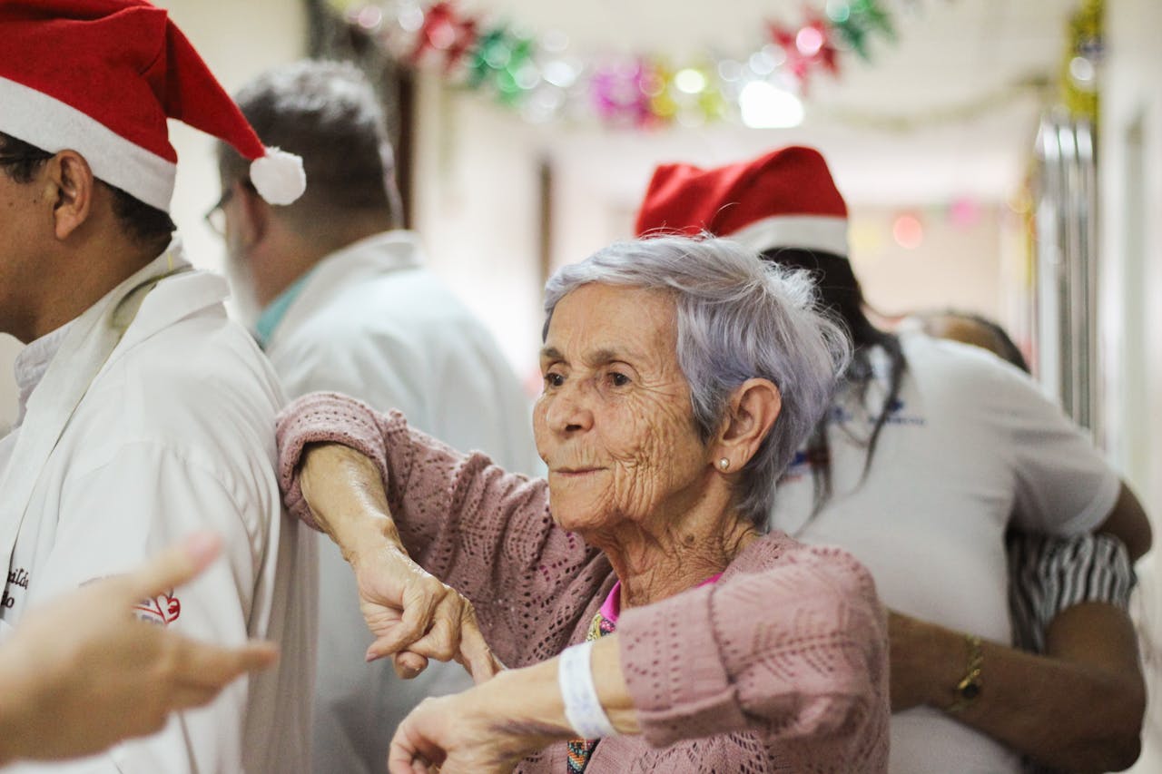 Home A joyful elderly woman in a festive setting, surrounded by people wearing Santa hats.